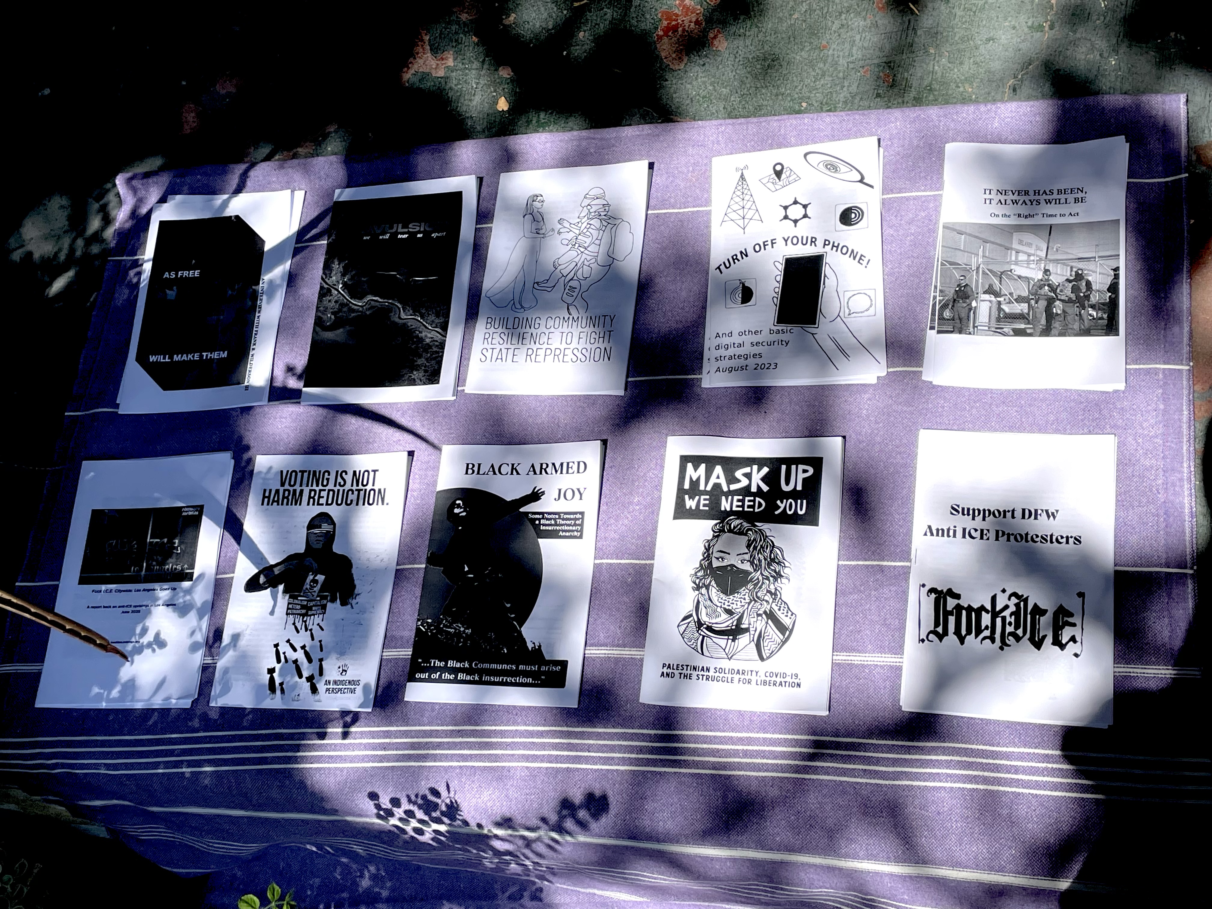 Photo of several black and white zines on a purple blanket. Titles include: Black Armed Joy; Mask Up: We Need You; Support DFW Anti-ICE Protesters; Voting is Not Harm Reduction; and Turn Off Your Phone!